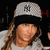 A pixelated close-up of a person wearing a light gray baseball cap with a dark emblem, long brown hair, and a light-colored shirt against a dark background.
