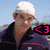 A person wearing a white cap and dark jacket with red stripes stands against a bright blue sky background, with a pink '<3' symbol visible in one frame.