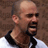 Static 48x48 pixel icon of a bald man with dark sunglasses and beard, wearing black shirt over white collar, against brown background.