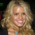 Pixelated close-up of a person with long, wavy blonde hair, wearing a light-colored top with a blue accent, against a dark background.