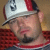 Static pixelated image of a person wearing a white and red cap with a black brim, visible facial features including eyes, nose, mouth, and beard stubble, and a red shirt against a brown background.