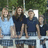 Four figures stand side-by-side on a light-colored platform with green foliage in the background; two wear dark blue shirts, one wears white, and one wears light blue.