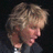 Pixelated close-up of a person with light brown curly hair, wearing dark clothing and holding a microphone near their mouth.