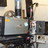 Pixelated static image showing a dark mechanical object with metallic and brown components against a light background.