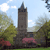 A static pixelated image shows a tall stone tower with a spire, surrounded by green trees and pink flowering bushes under a light blue sky with white clouds.
