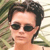 Pixelated close-up of a person with short dark hair wearing dark sunglasses against a blurred green and beige background.