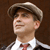 Pixelated image of a person wearing a light-colored flat cap, white shirt, and red tie, with a brown background.