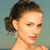 Pixelated close-up of a person with dark curly hair tied in a low bun, light skin, and a neutral expression against a muted teal background.