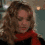 A four- shows a person with curly brown hair wearing a red scarf and dark top against a blurred background with lights