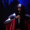 Animated sequence of a person in dark clothing with red accents holding a microphone, performing on a stage with blue lighting.