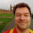 Pixelated static image of a person with short brown hair wearing red and yellow clothing, standing outdoors near green grass and wooden structures.