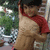 A shirtless man with a red cloth draped over his shoulder stands outdoors, holding an object near his chest.