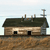 Pixelated static image of a small wooden house with dark roof, two black windows, and multiple chimneys against light sky background.
