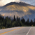Pixelated landscape showing a road with yellow lines, dark trees, and a sky with layered clouds in blue, orange, and gray tones.