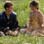 Two pixelated figures sitting on grass, one in dark clothing and the other shirtless with brown hair, against a green background.