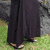 A woman with long red hair wears a black sleeveless dress, standing against a stone wall with grass visible at the bottom left.