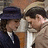 Two figures in formal attire embrace; one wears a dark blue hat and white shirt, the other has light brown hair and a white tie.