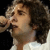 Pixelated close-up of a person with curly dark hair holding a microphone near their mouth, wearing a light-colored top against a dark background.