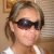 Pixelated close-up of a person with light brown hair tied back, wearing dark sunglasses and a white top against a blurred background.