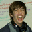 A pixelated image of a person with brown hair and headphones around their neck, wearing a dark jacket over a gray shirt, against a light background.