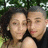 Two people pose closely together against a blurred green background with visible foliage, one with dark curly hair and the other with short gray hair.
