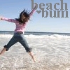 A person in a pink striped shirt and dark pants jumps on a beach with arms and legs spread wide, against a backdrop of ocean waves and clear sky.