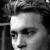 Black-and-white pixelated portrait of a person with dark hair, visible eye, nose, and mouth, set against a dark background with light horizontal lines.
