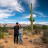 Two people stand side-by-side in a desert landscape with a tall saguaro cactus under a partly cloudy blue sky.