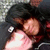 Two people in close embrace, one with dark hair resting head on shoulder of another in dark jacket with white button, against blurred background.