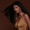 A woman with long dark hair wears a sleeveless light brown top against a solid brown background.