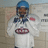 A person in a white sports uniform with blue and red accents stands against a blurred background, wearing a blue helmet.