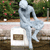 A pixelated silver statue of a seated figure with a rounded head and outstretched arm, positioned on a white pedestal with a brown rectangular plaque, against a blurred green and brown background.