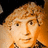 Pixelated static image of a person with light brown curly hair, neutral expression, and warm-toned background.