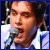 Pixelated static image of a person holding a microphone against a dark background with light reflections, wearing a white shirt and dark jacket.