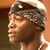 Pixelated close-up of a person's face with dark hair and light skin, wearing a white headband or cap against an orange-brown background.