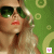 A pixelated image of a woman with blonde hair and sunglasses against a green background with light yellow circles, featuring a small pink or purple icon in the corner.