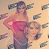 Two women pose together against a backdrop with repeated dark blue text; one wears a strapless red dress, the other a light-colored top.