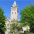 Pixelated static image of a tall stone tower with a spire, surrounded by green trees under a clear blue sky.