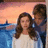 Two figures stand side-by-side against a sunset sky with blue water below; the figure on the left has long brown hair and wears white, while the right figure has short dark hair in a blue shirt.