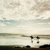 Silhouetted figures holding surfboards stand on a beach under a cloudy sky with layered clouds and calm water.