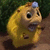Pixelated yellow creature with large black eyes, a small blue gem on its head, and an open mouth holding a dark object against a green background.