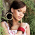 A pixelated image of a person with dark hair tied back, wearing a white sleeveless top with a red accent, holding a large black camera against a green background.