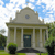 A pixelated image of a classical building with columns, a central door, and a circular window under a blue sky with scattered clouds.