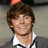 Pixelated portrait of a person with brown hair wearing a white collared shirt and dark jacket, smiling against a muted background.