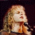 Two side-by-side pixelated figures with blonde hair; left figure holds microphone, right figure has neutral expression against striped background.
