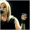 A person with long blonde hair wearing a black sleeveless top holds a microphone close to their mouth against a dark background.