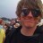 Pixelated image of a person with brown hair wearing dark sunglasses and a dark shirt, standing outdoors near water.