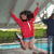 Pixelated image of a person in red long-sleeve shirt and light shorts with arms raised high, standing on a platform near water.