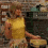 A person in a yellow sleeveless top and light shorts holds a round yellow object in a kitchen setting with dark cabinets and a tiled wall.