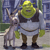 Green ogre in white tunic and brown boots stands beside gray donkey with white mane, both facing forward against blurred blue-green background.